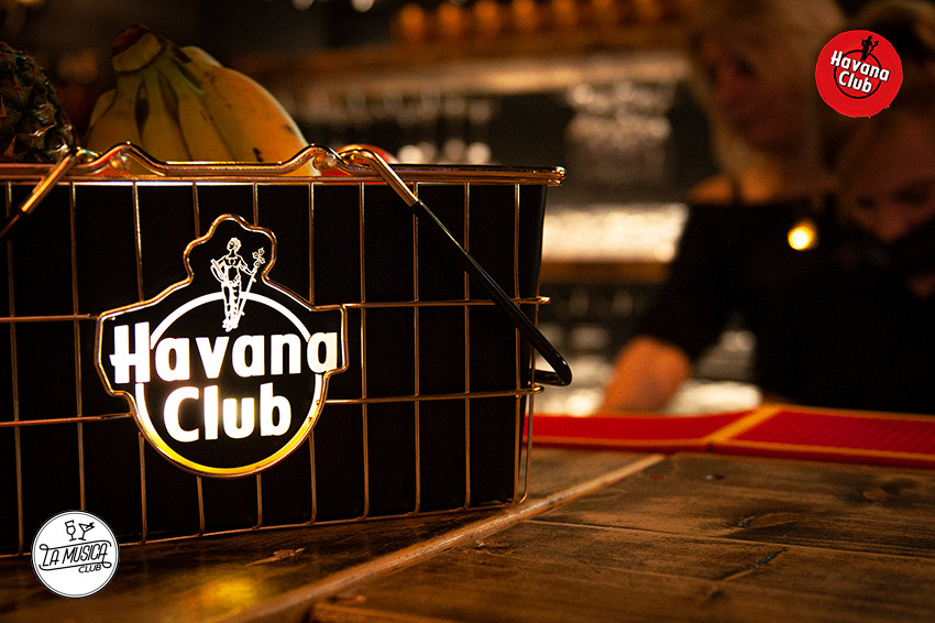 Metalowy koszyk z logo Havana Club na drewnianym barze, w tle ananas, banany i barman